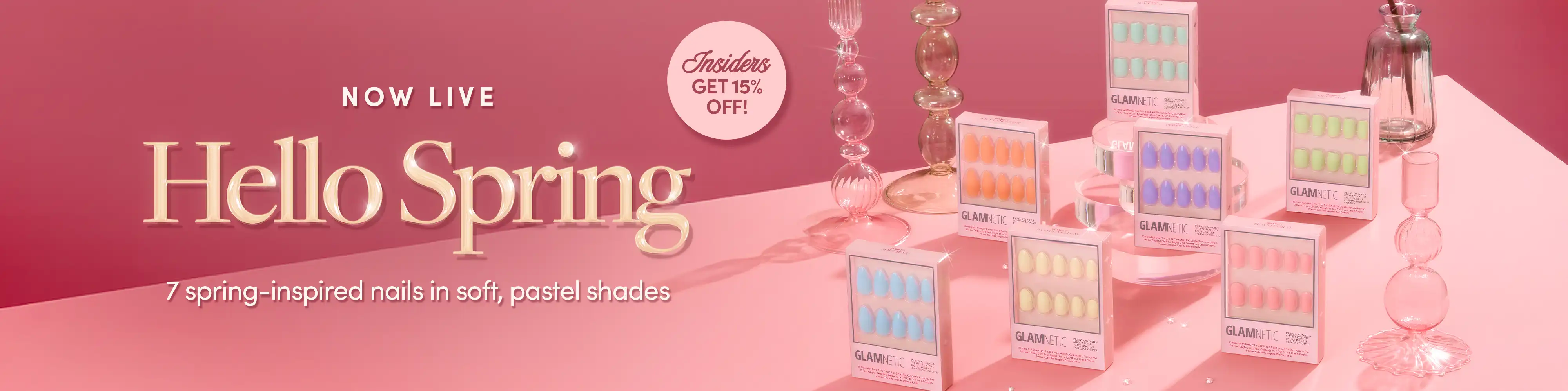 Pastel-colored press-on nail kits displayed on a pink surface with glass candle holders. Text reads: Hello Spring. 7 spring-inspired nails in soft, pastel shades. Exclusive! Get 15% off!.