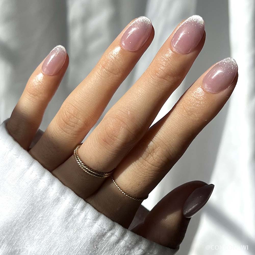 A hand with neatly manicured short oval nails in glamnetic’s Fairy Dust, featuring a soft pink base and white gradient tips, wears two delicate gold rings and is partially covered by a white long-sleeve shirt. - Product image
