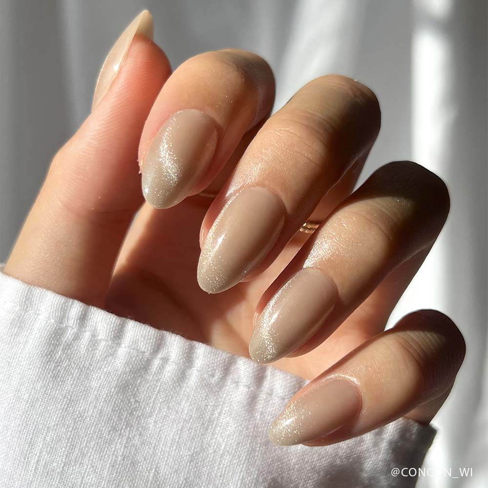 A hand with short almond-shaped nails painted in glamnetics Honey Bun, a nude matte polish with subtle shimmer and a velvet finish, shown in natural sunlight. The person wears a white long-sleeve top. - Product image