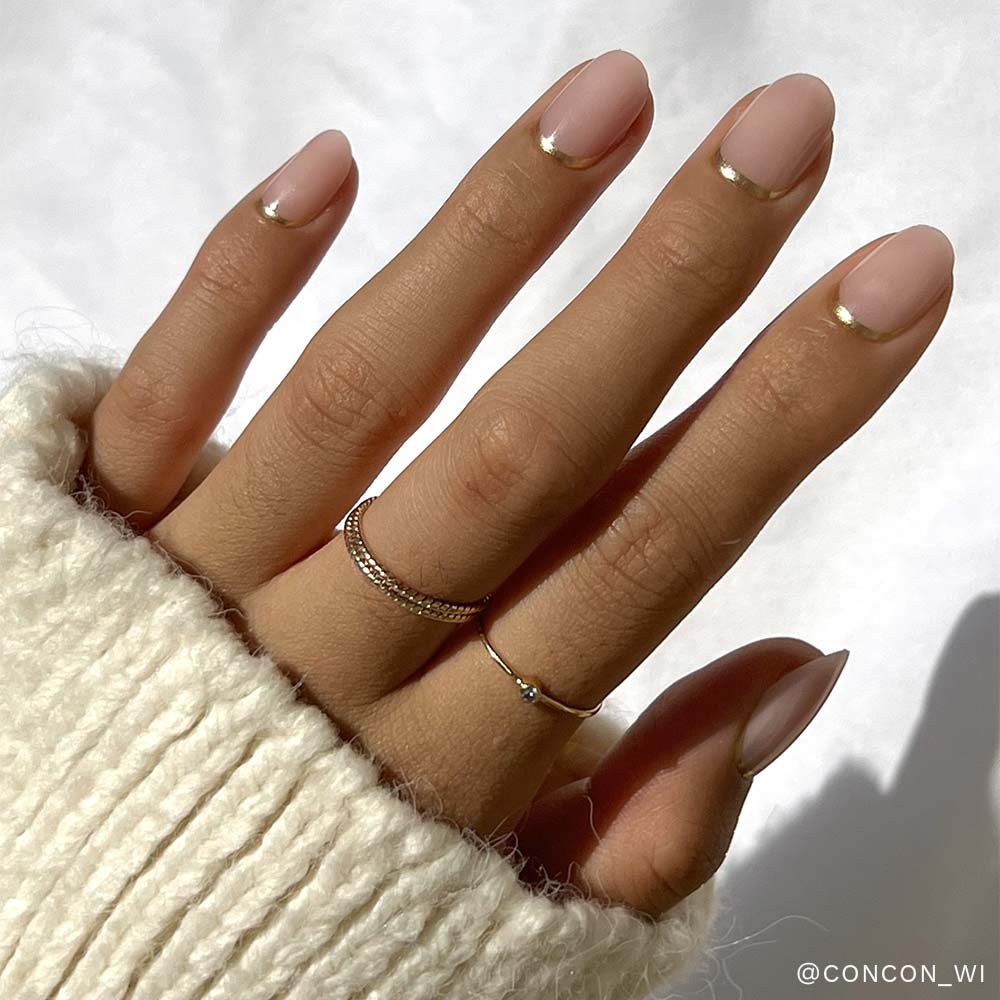 A hand with a nude manicure and thin metallic gold tips from glamnetic's Nail Lover's Gift Set is shown, accessorized with delicate rings and a cozy cream knit sweater, sunlight highlighting the fingers. - Product image