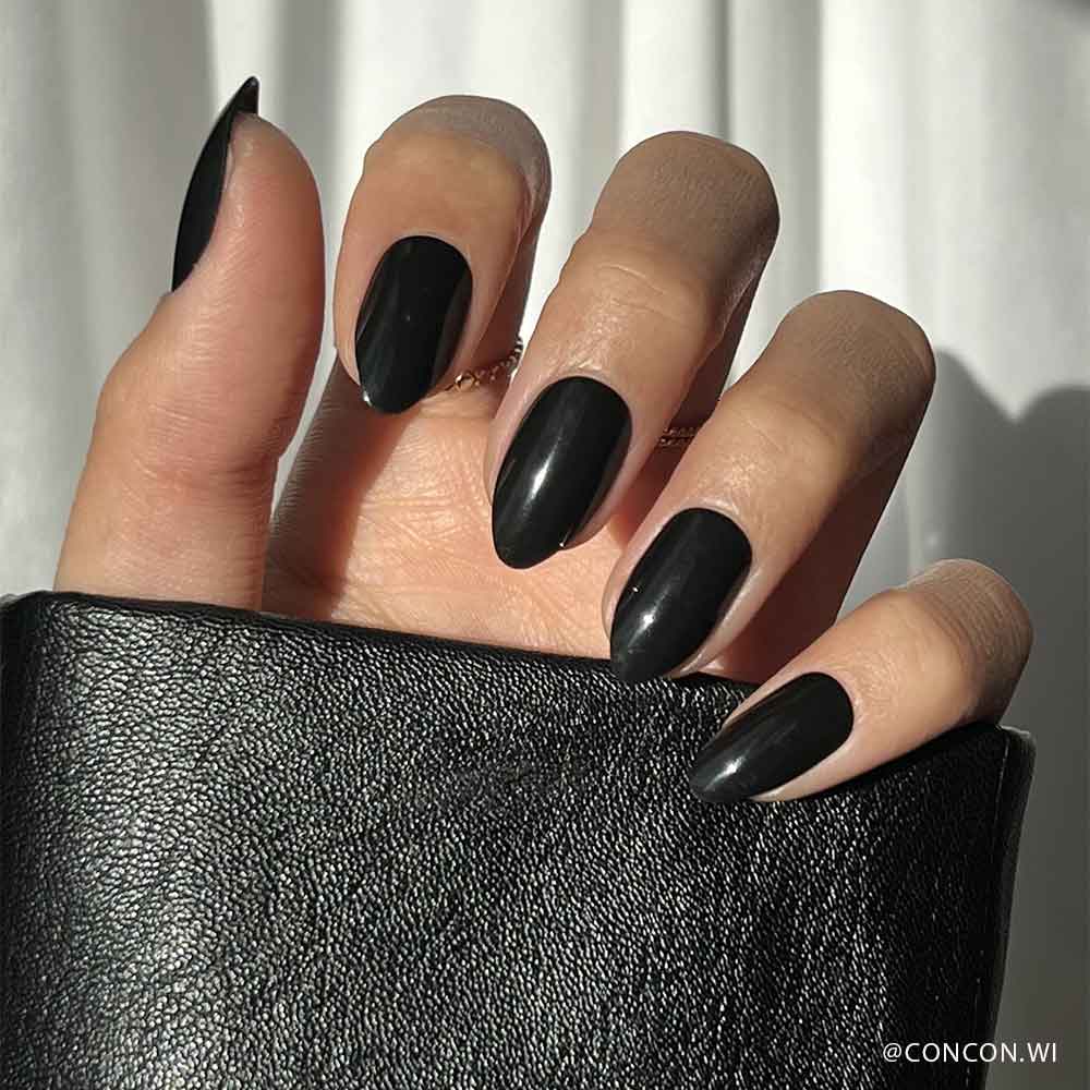 A hand with long, glossy black glamnetic Boba vegan cruelty-free nails holds a textured black leather item in bright lighting, with a white curtain in the background. - Product image