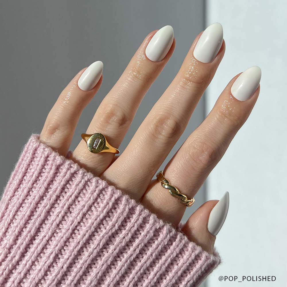 A hand with almond-shaped, glossy white Angel vegan press-on nails by glamnetic, adorned with two gold rings, is shown against a neutral background. The person wears a pink ribbed sweater with the sleeve partially covering their fingers. - Product image