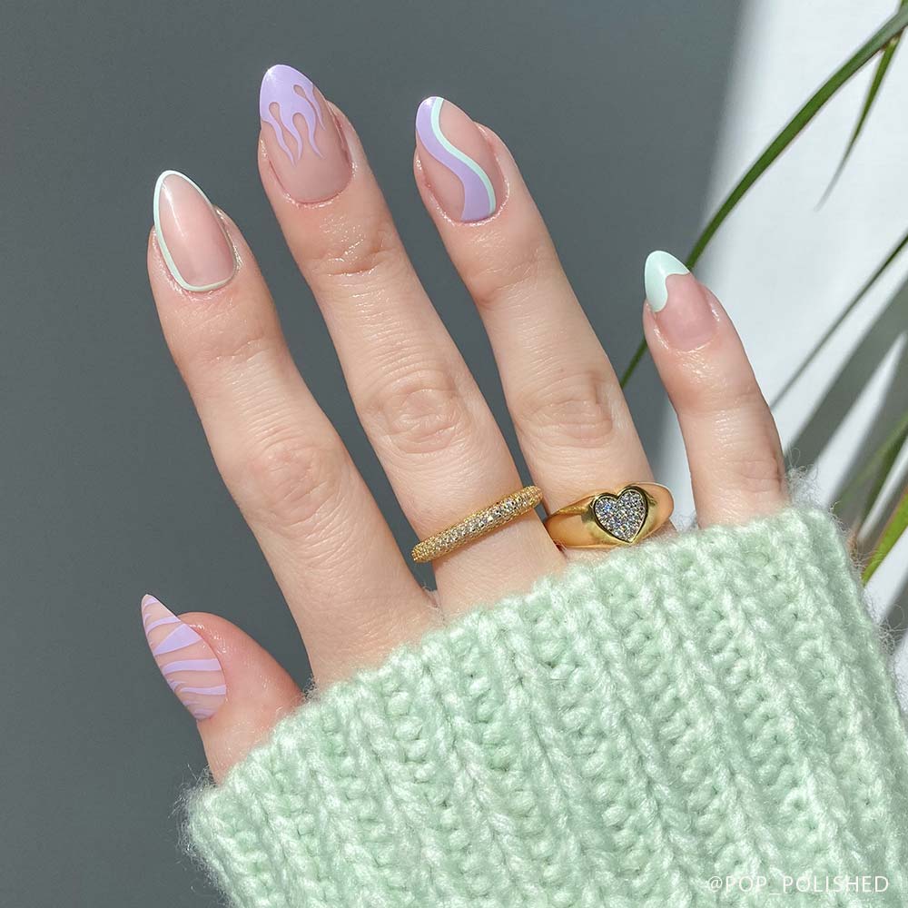 A hand with long, almond-shaped glamnetic Wild Card press-on nails in semi-transparent pastel waves, swirls, and flames—pink, purple, and mint. Two gold rings and a mint green knit sweater complete the chic look. - Product image