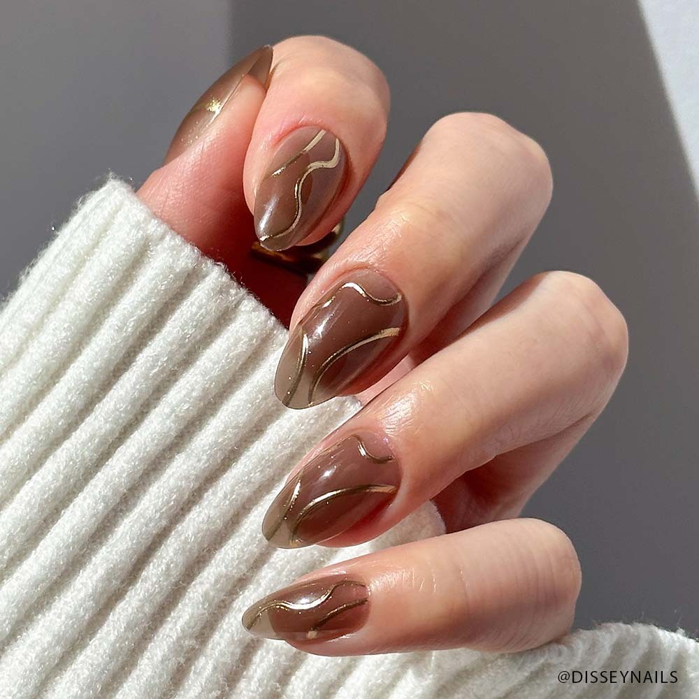 A hand with long, almond-shaped nails from glamnetic’s Golden Cup™ set, featuring a glossy brown base and gold wavy lines. The person is wearing a white ribbed sweater. - Product image