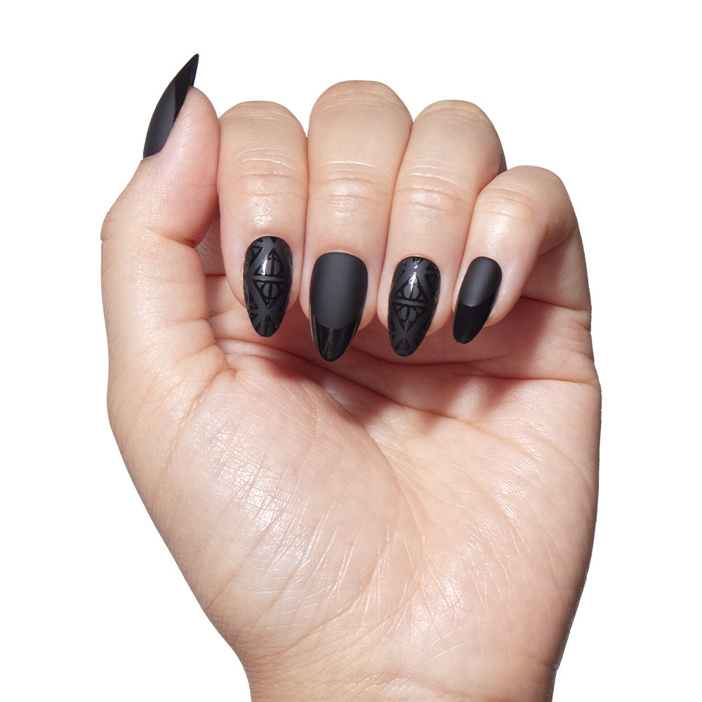 A hand displays long, almond-shaped nails painted matte black with glossy black Deathly Hallows™ geometric designs by glamnetic on two nails, all set against a plain white background. - Product image
