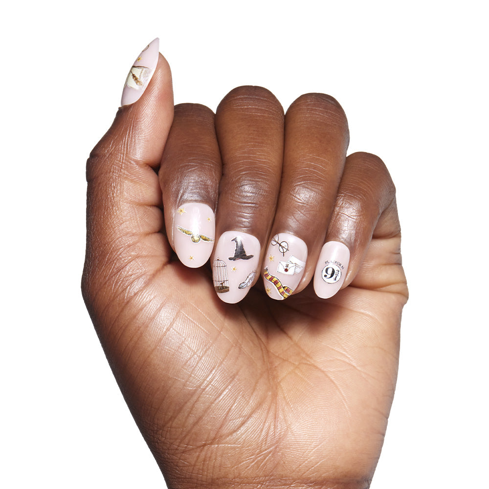 A hand with light pink glamnetic Hogwarts™ nail polish showcases intricate Harry Potter designs—broomstick, glasses, witch hat, and 9 3/4—making the art pop against a plain white background. - Product image