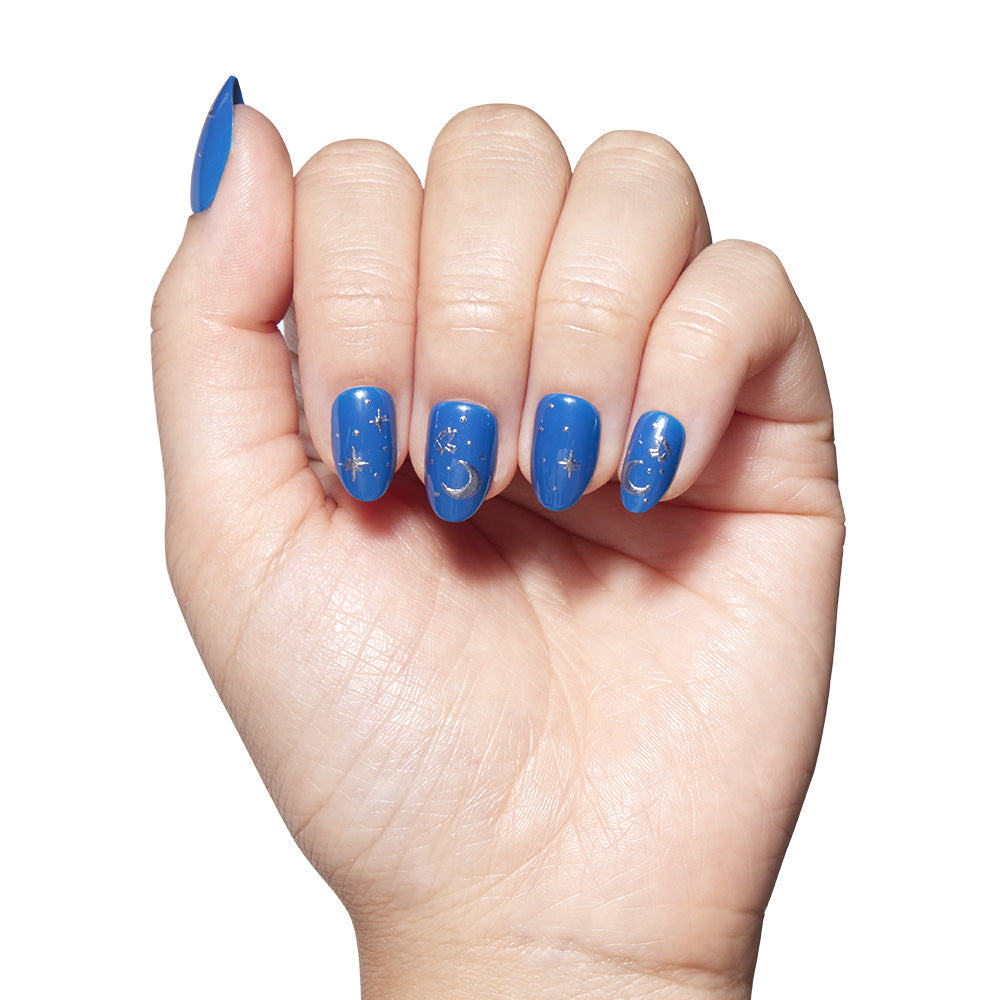 A hand with short oval nails painted blue and decorated with gold stars, moons, and silver foil—part of the glamnetic Luna Lovegood™ set—is displayed against a white background. - Product image
