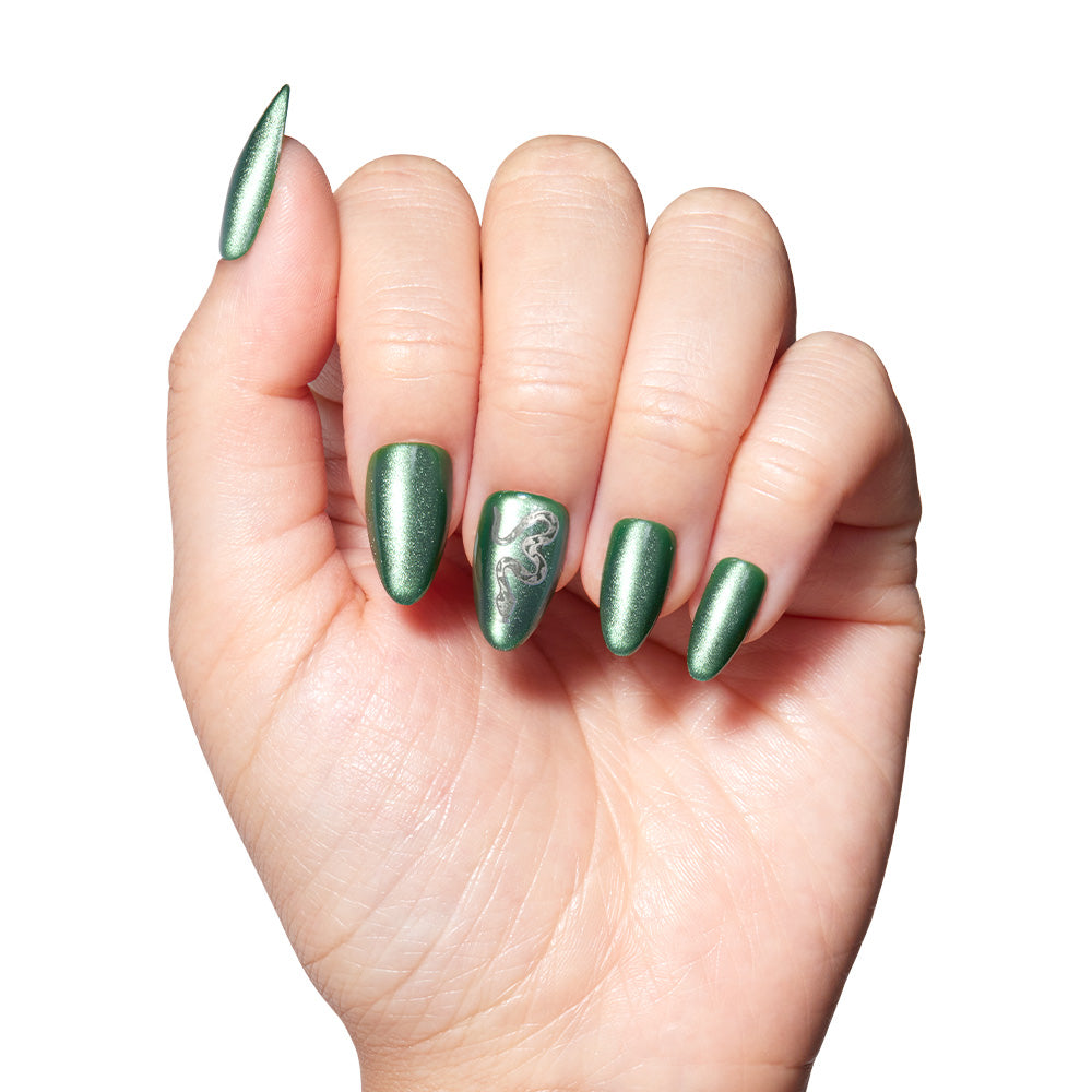 A hand displays long, almond-shaped, emerald green metallic nails from the glamnetic Slytherin™ set. The ring fingernail features a silver swirl design, and the sleek set pops against a plain white background. - Product image