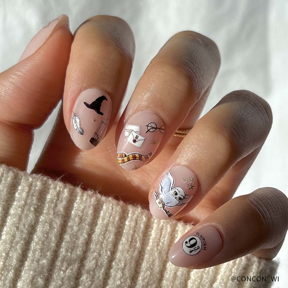 A hand with light pink glamnetic Hogwarts™ nail polish featuring a wizard hat, scarf, Hedwig, Hogwarts letter, and glasses. The person is wearing a beige knit sweater. - Product image