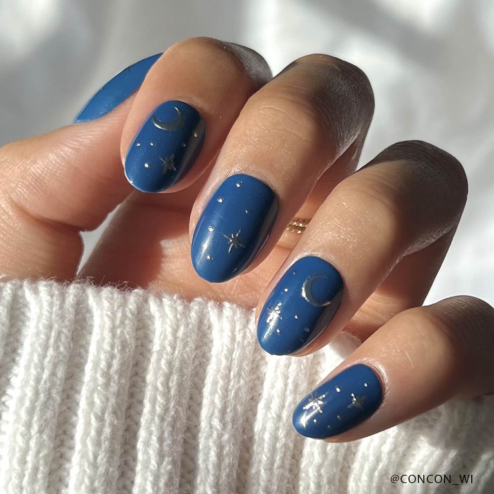 Short oval nails with blue matte polish, minimal gold crescent moons and stars, and tiny silver foil accents are styled against a white knit sweater—an enchanting look created using the Luna Lovegood™ set by glamnetic. - Product image