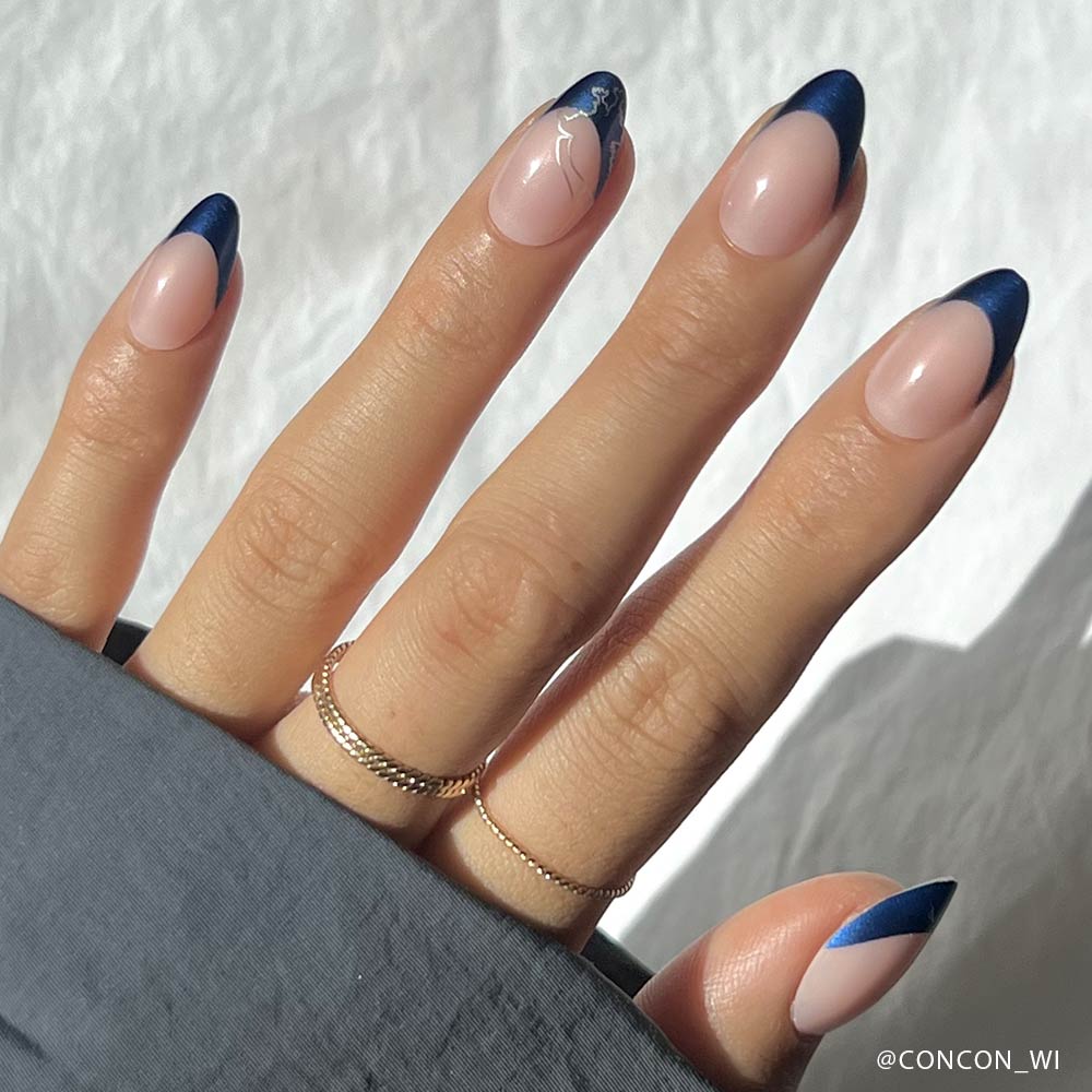 A hand with short almond-shaped nails wears glamnetics Ravenclaw™: a nude base, sapphire blue chrome French tips, and a delicate white abstract line on one nail. Gold rings and a gray sleeve complete the look. - Product image