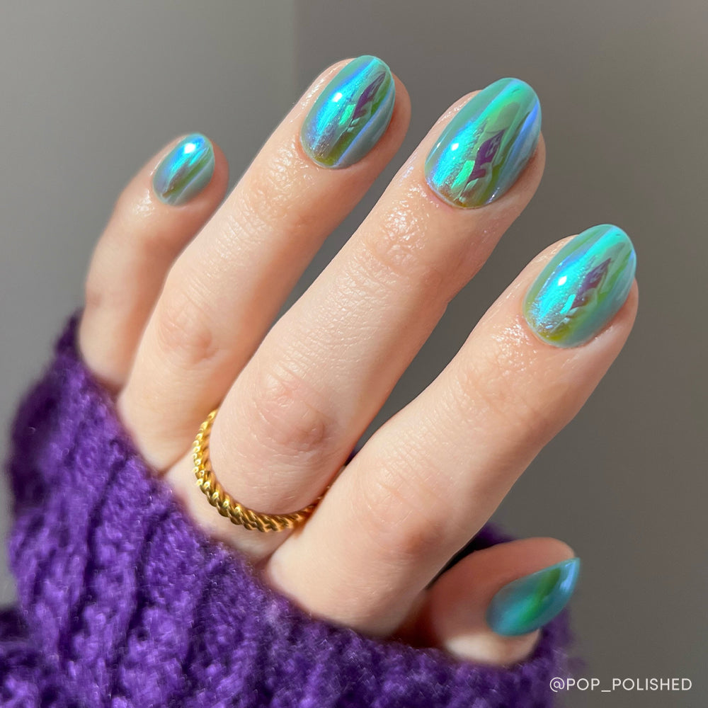 A hand with short oval-shaped nails painted in Siren by glamnetic—a shiny, holographic teal and green chrome—pairs perfectly with a gold twist ring and purple knit sweater. - Product image