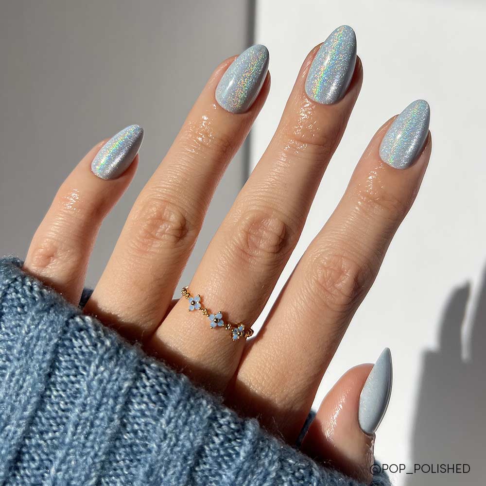 The Frozen nail set by glamnetic features short almond-shaped nails in an icy-blue shade. The hand is accessorized with a delicate gold ring with blue stones and partially covered by a blue knit sweater. - Product image