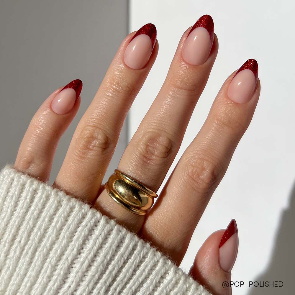A hand with short almond-shaped nails features glamnetic’s Red Affair press-ons, showcasing a nude base with dark red glitter French tips. The person wears a chunky gold ring and a white ribbed sweater. - Product image