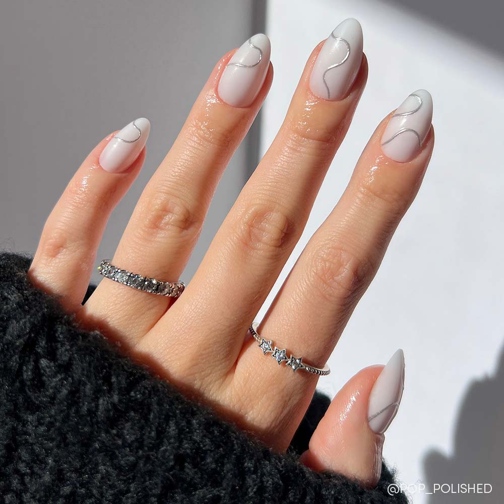 A hand shows off Silent Night by glamnetic, a short almond nail set with a white minimalist swirl. The person wears silver star and gem rings plus a black textured sweater, all softly lit for a chic winter vibe. - Product image