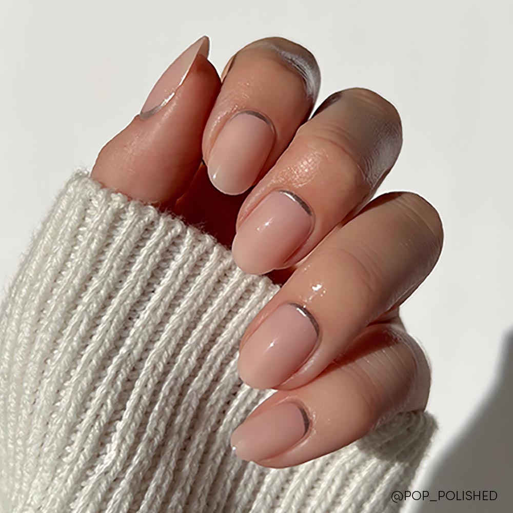 A hand with short, round nails showcases glamnetic’s Silver Belle—a minimalist nude polish with thin silver lines at the cuticles. The person wears a cream-colored ribbed sweater. - Product image