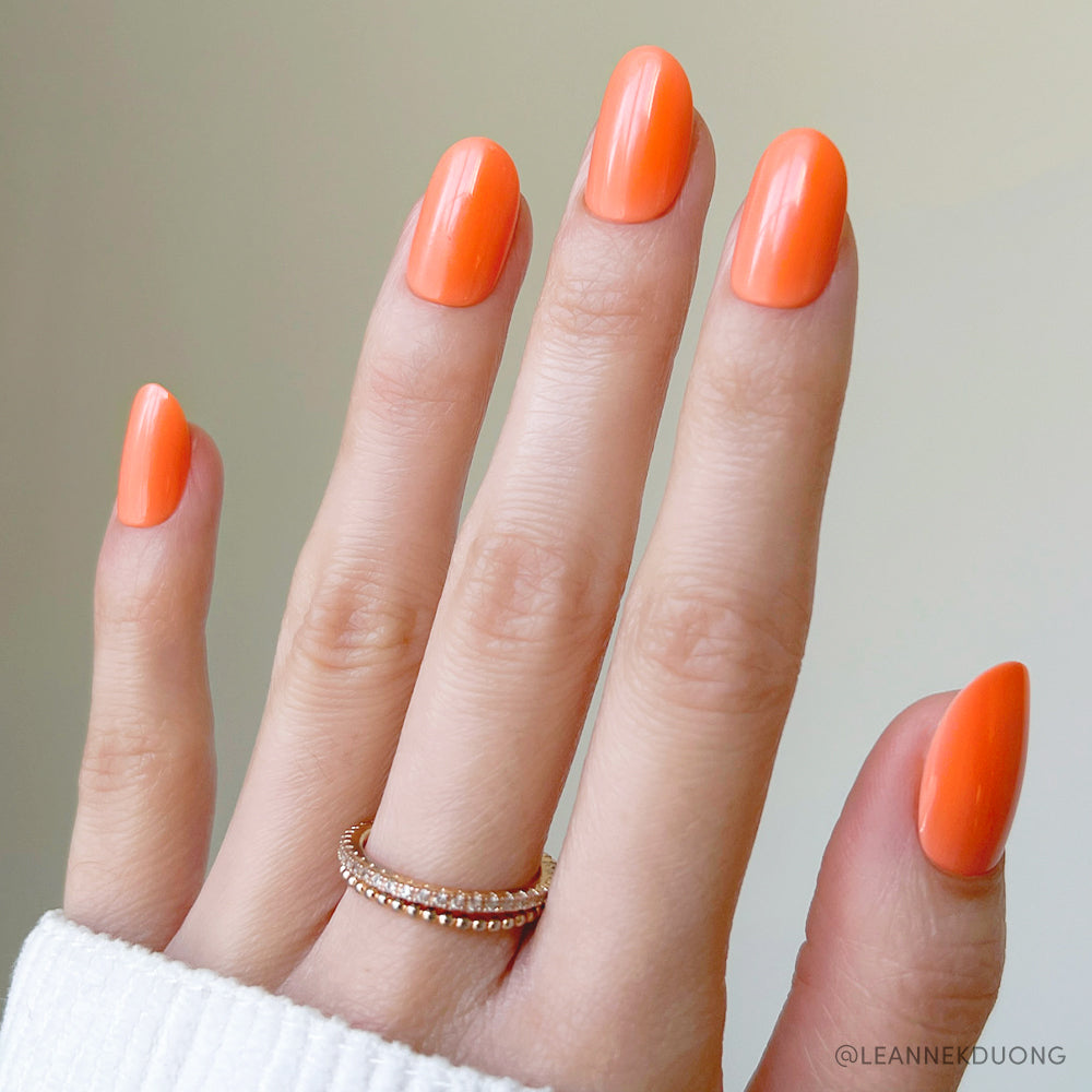A hand with long, almond-shaped Apricot press-on nails by glamnetic is shown in vibrant orange. Two gold rings accent the ring finger, a white sleeve covers the wrist, and the background is softly blurred. - Product image