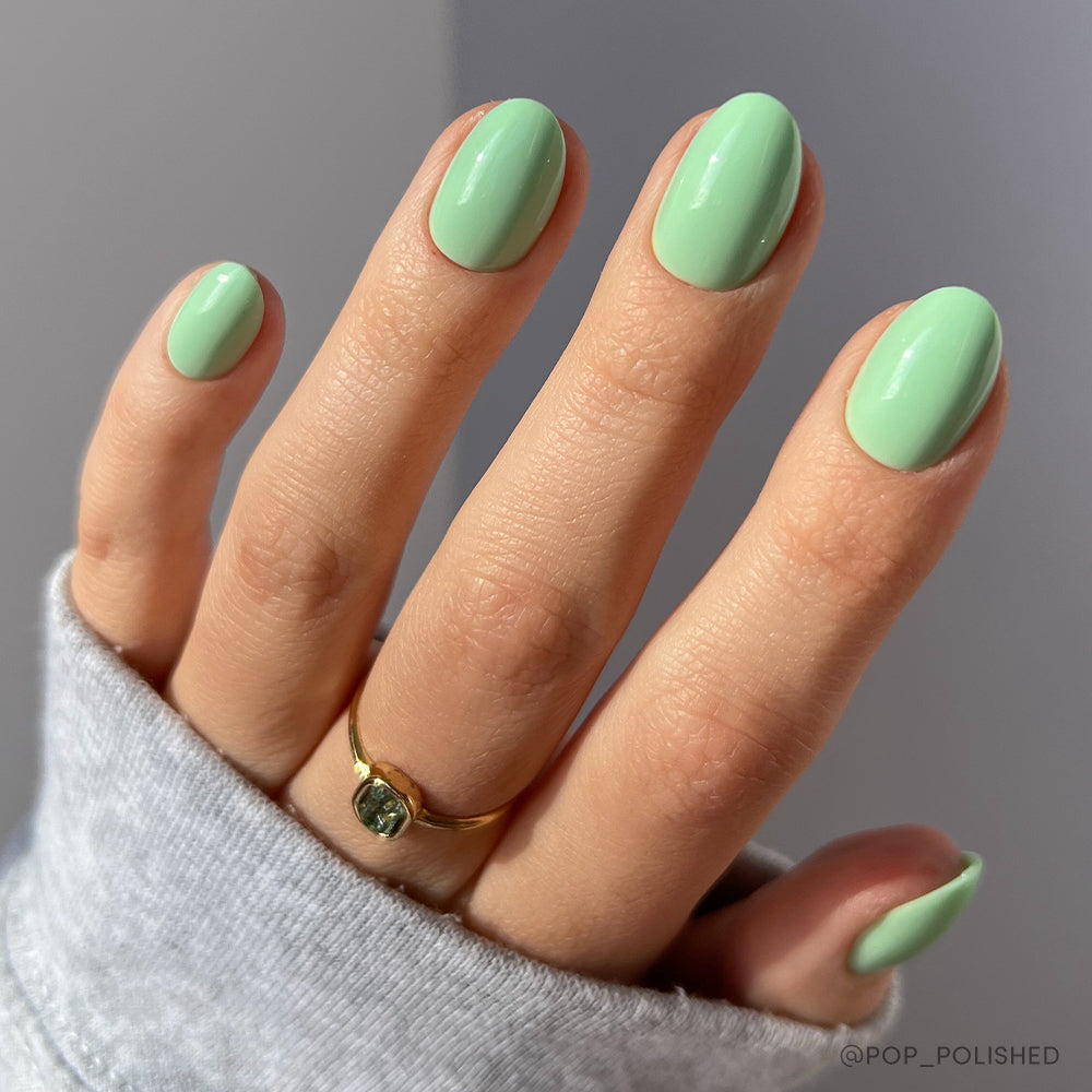 A hand with short oval-shaped nails painted in glamnetic’s Mint Green set, glossy finish, wears a gold ring with a green stone and is partially covered by a light gray sleeve. - Product image