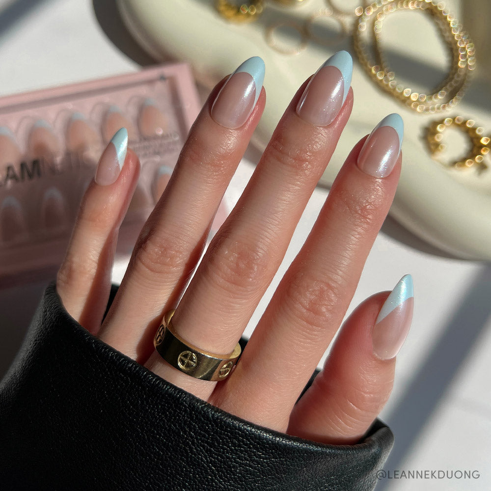 A hand with long, almond-shaped nails in glamnetic’s Blueberry Icing set—baby blue French tips and a glazed finish—wears a gold ring, is partly covered by a black sleeve, with the Blueberry Icing box and gold jewelry in the background. - Product image