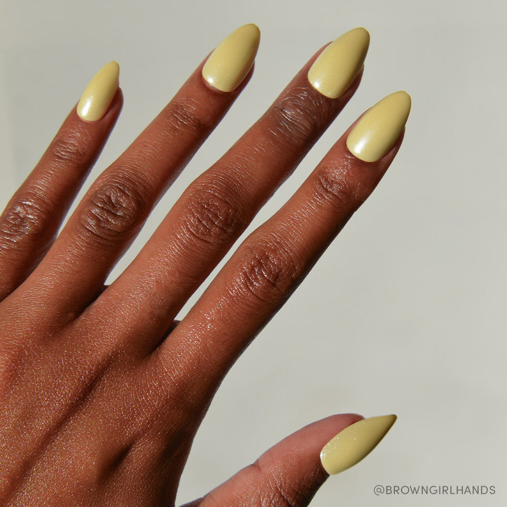 A close-up of a hand with medium-brown skin and long, almond-shaped nails painted in Yellow Pop by glamnetic. The plain, light background highlights the soft charm of this pastel yellow nail set. - Product image