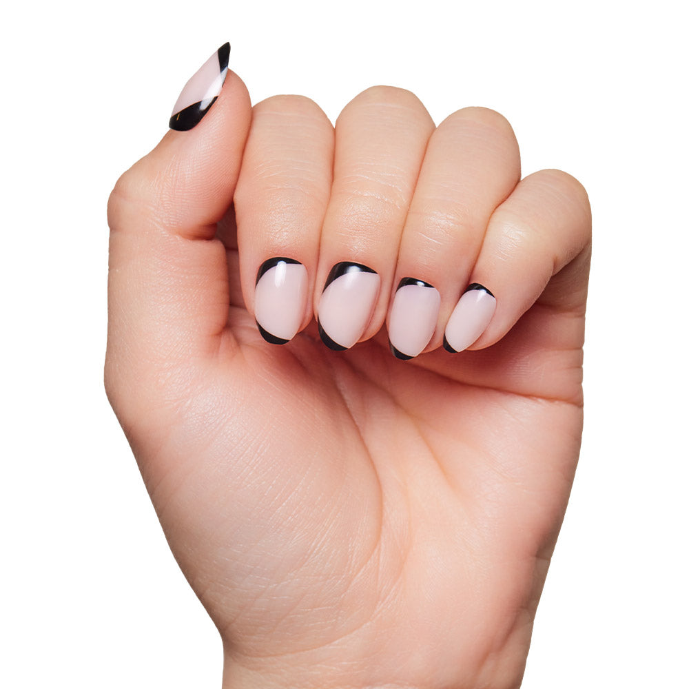 A hand with short, almond-shaped nails featuring a nude base and black abstract tips from the glamnetic NYC press-on set, shown against a white background. - Product image