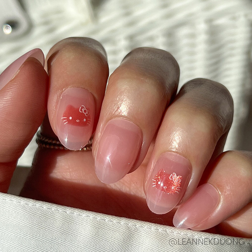 A hand with oval-shaped nails from the glamnetic Hello Kitty® set, painted sheer pink. Two nails show white Hello Kitty faces—one on a coral pink accent—all vegan, cruelty-free, and shown in sunlight over a woven background.