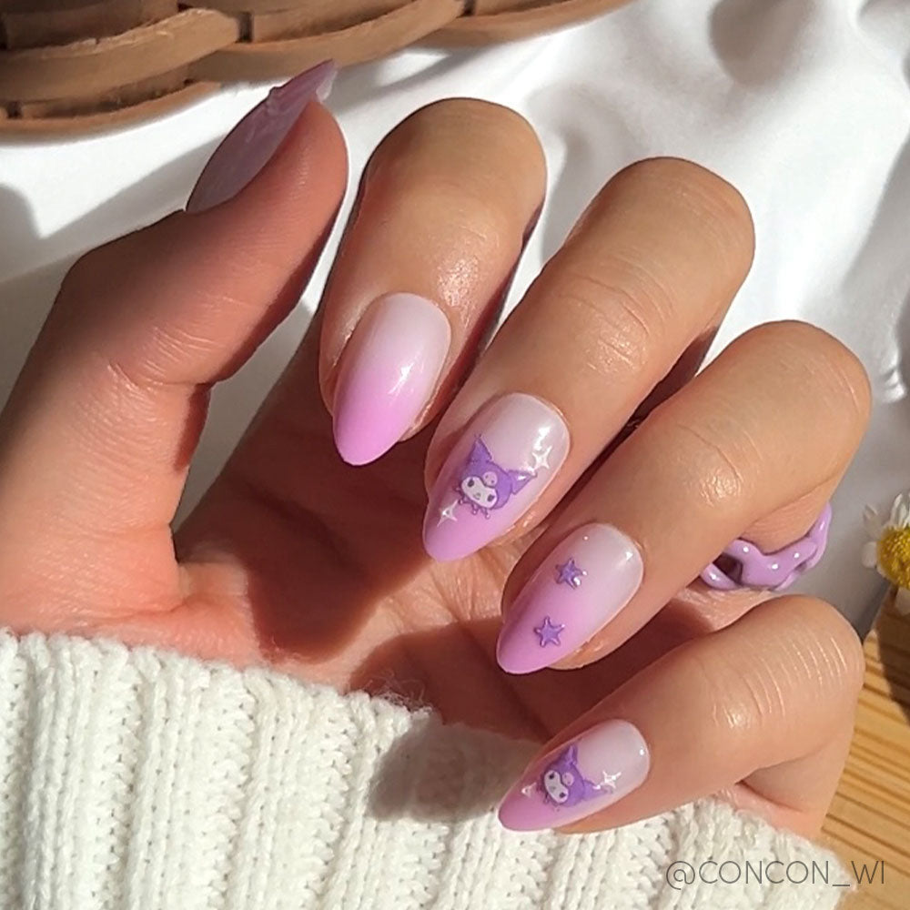 A hand with long, almond-shaped nails painted in a pink-to-lavender gradient featuring purple Kuromi™ designs, stars, 3D accents, and a purple ring. The person wears a white sweater. Vegan & Cruelty-free by glamnetic.