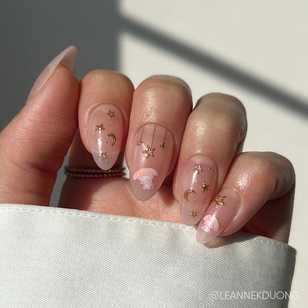 A hand wears glamnetic’s My Melody™ short almond nails with a soft pink base, gold star and moon decals, cloud accents, and subtle My Melody art. Partially covered by a white sleeve, the nails shine in natural sunlight.