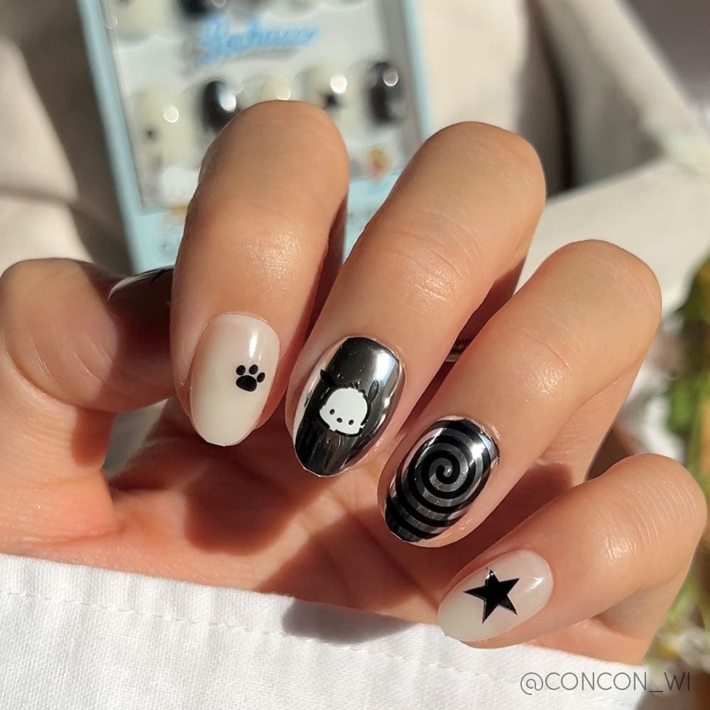 A hand displays chic press-on nails from the Pochacco™ set: a black paw print, Pochacco character face, black and white spiral, and a black star on nude or white nails.