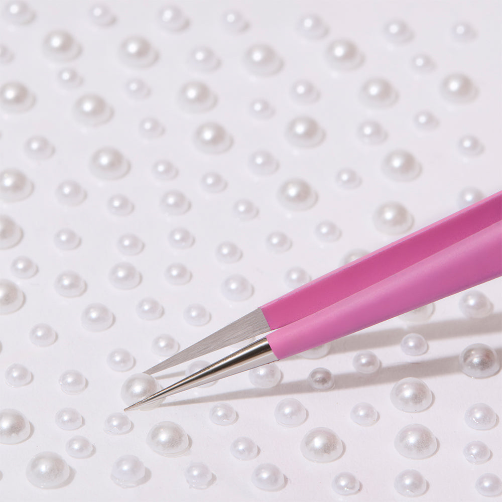 A pair of glamnetic Nail Decal Tweezers with an ultra-precise design picks up a small, round, white pearl from scattered pearls on a white surface. - Product image