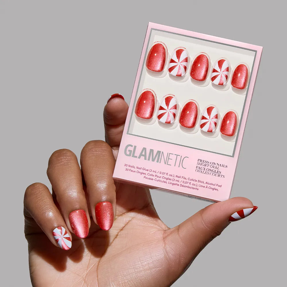 A hand with red and white peppermint swirl nails holds a pink glamnetic box, showcasing the Candy Crush holiday nail set—10 matching red and white designs—against a neutral gray background.