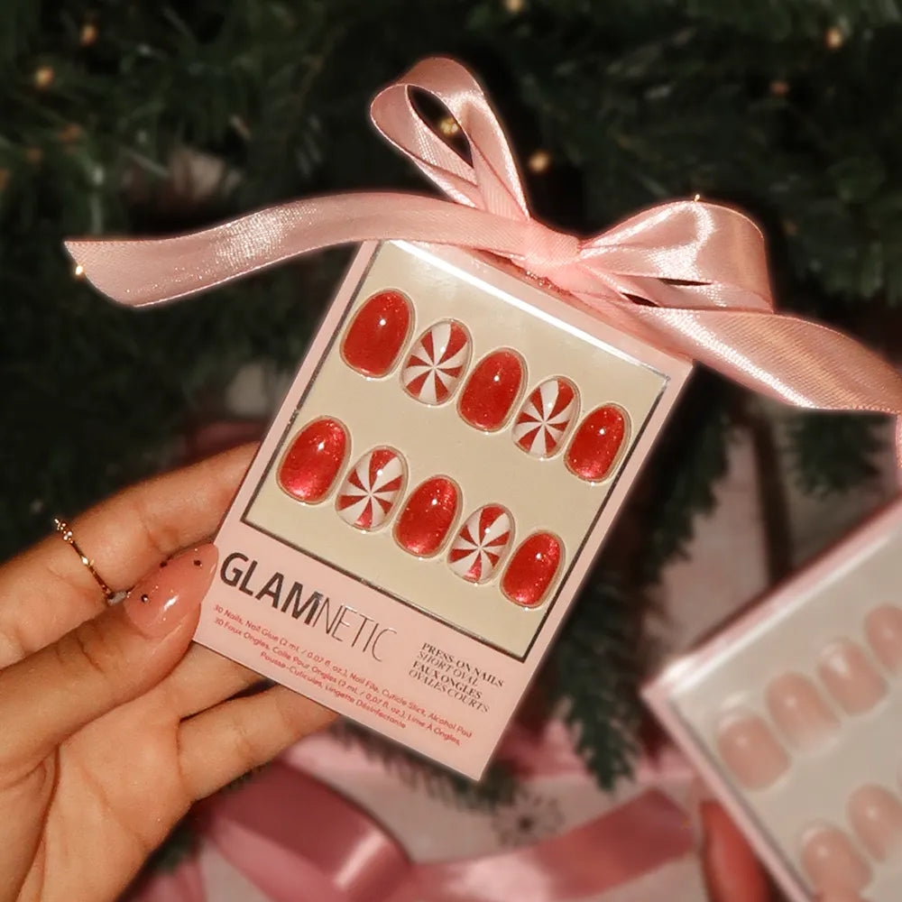 A hand holds a box of glamnetic's Candy Crush press-on nails, featuring peppermint swirl designs with a pink ribbon—perfect for the holidays, with a blurred Christmas tree in the background. - Product image