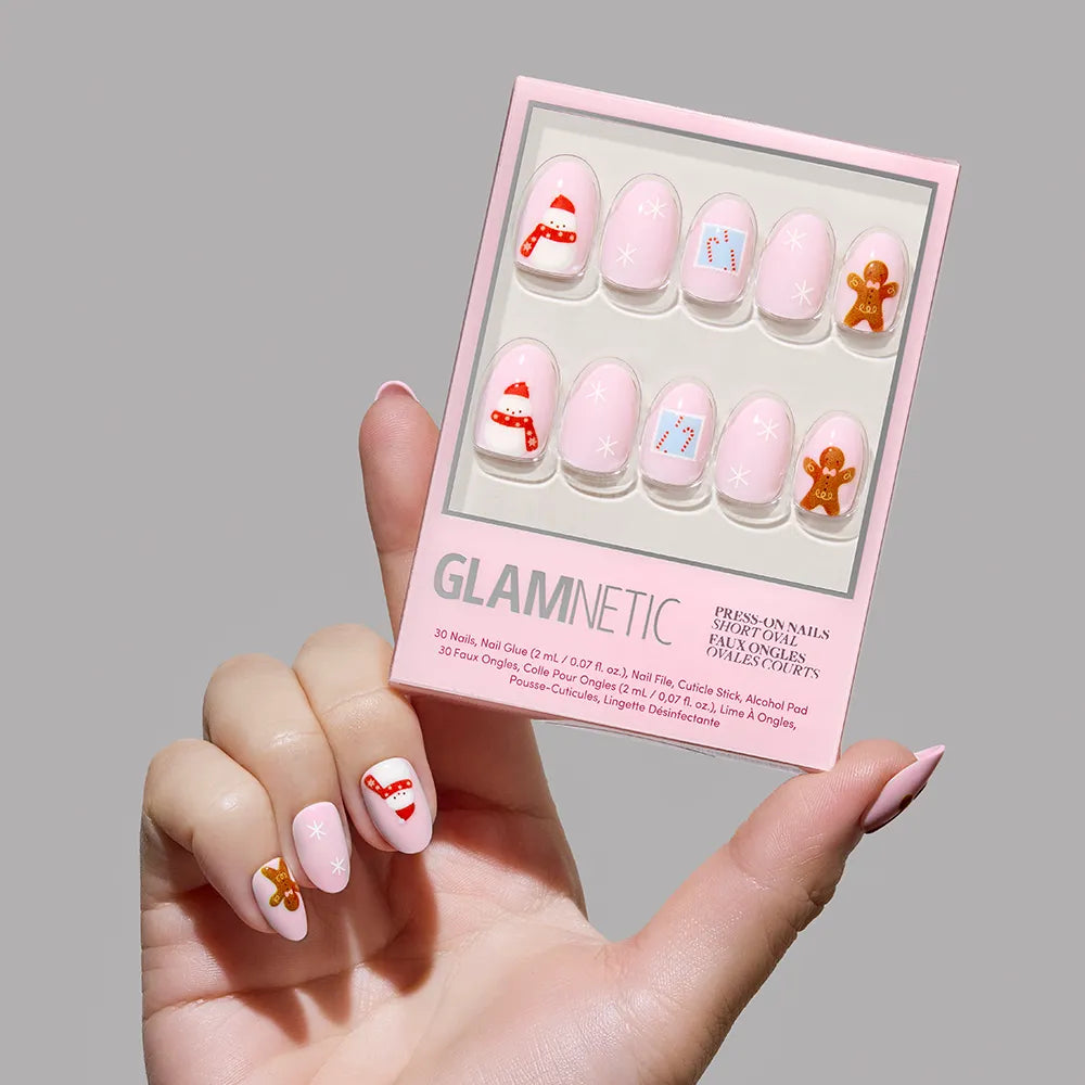 A hand holds a box of glamnetic Cocoa & Kisses short oval nails, a holiday set with festive designs—candy canes, gifts, snowflakes, and gingerbread men—on a light pink background. The wearer has matching themed nails.
