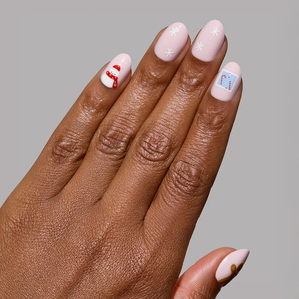 Short oval nails painted with glamnetic Cocoa & Kisses, a light pink polish, display festive designs like white snowflakes, a red scarf, a heart envelope, and a brown ornament on a plain gray background. - Product image
