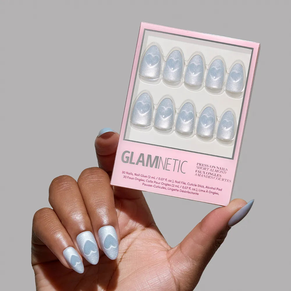 A hand with short almond-shaped, light blue nails with white hearts holds the glamnetic Cold Hearted set against a plain gray background.