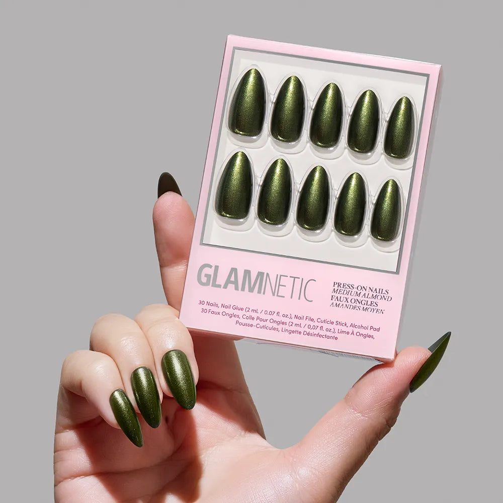 A hand with long, emerald almond nails holds a box of Cutie Pine green press-on nails labeled glamnetic against a neutral background.