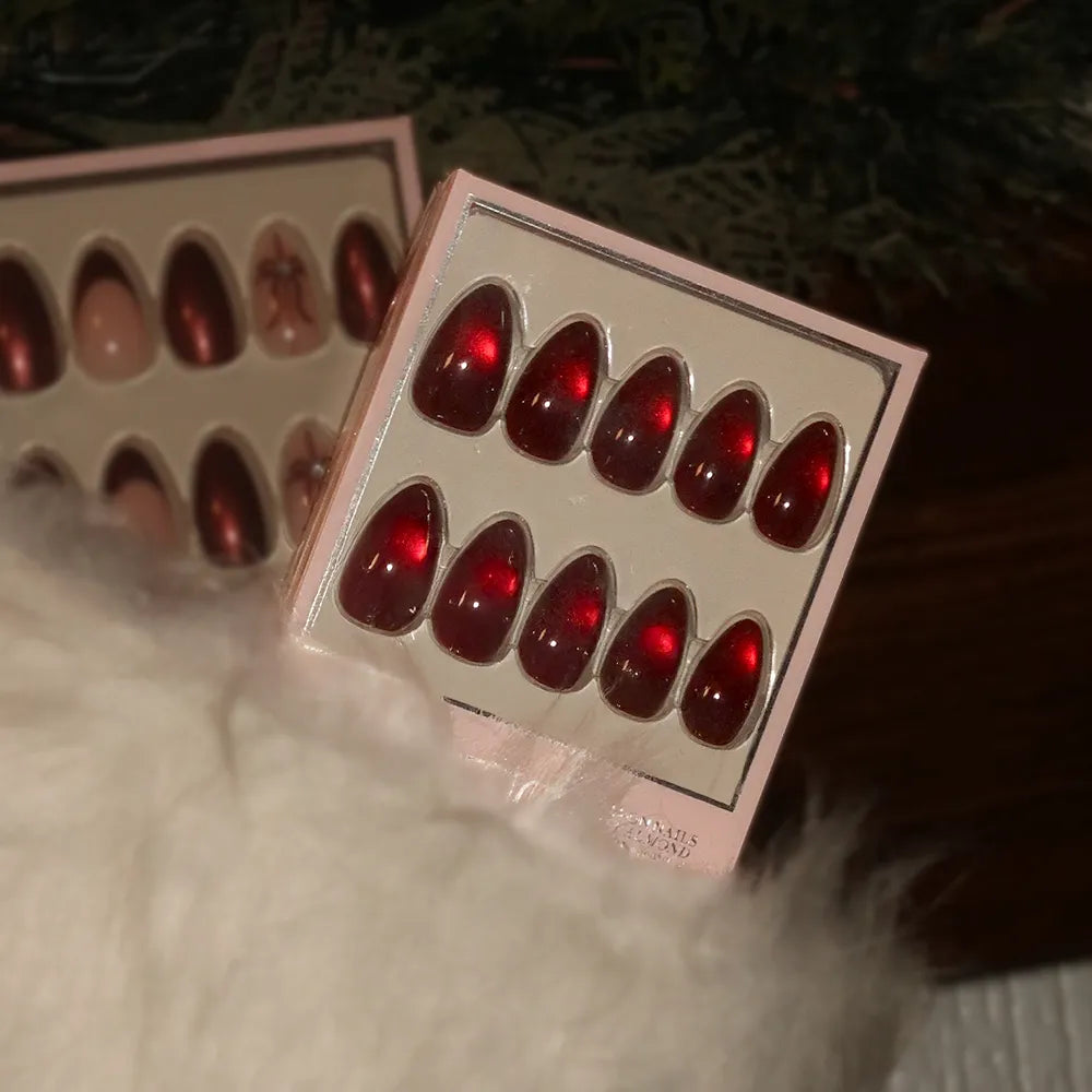 A box of glamnetic Frosted Cranberry glossy red press-on nails is shown partially tucked in a fluffy white stocking, with another festive nail set visible in the background. - Product image