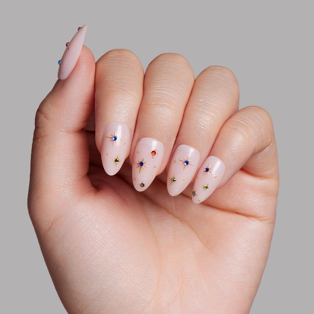 A hand displays long almond nails painted pale pink, each featuring colorful rhinestones and glamnetic North Star set-inspired starburst accents, shown against a plain gray background. - Product image