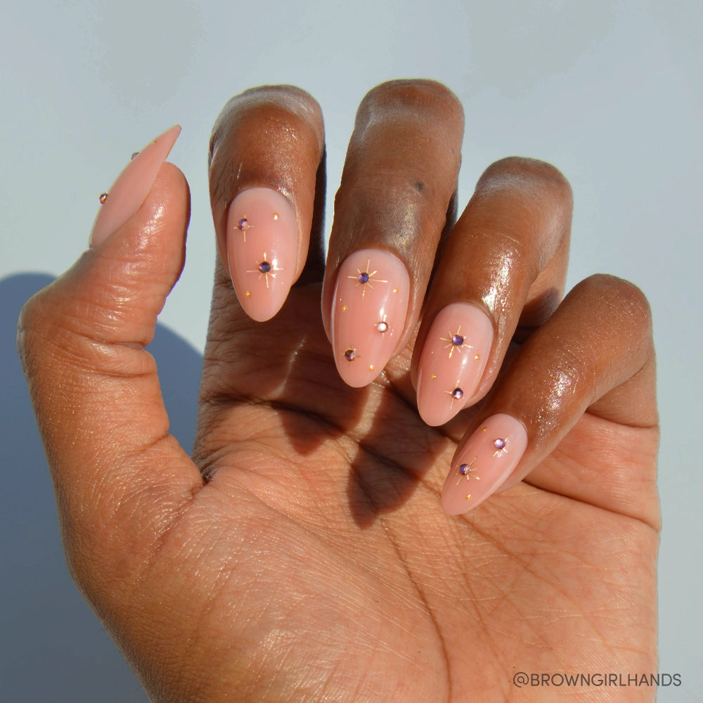 A hand with medium brown skin displays glamnetic's Pink Comet Nail Set—short almond nude pink nails featuring glossy finishes, gold foil stars, and gem accents—neatly manicured and shining in sunlight against a pale backdrop.