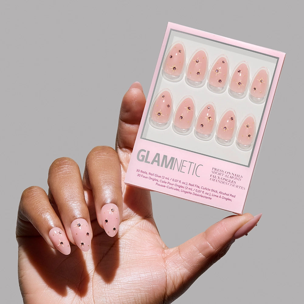 A hand with short almond-shaped Pink Comet press-ons by glamnetic, featuring light pink nails with black hearts, holds a matching glamnetic nail set in a pink box against a well-lit neutral background.