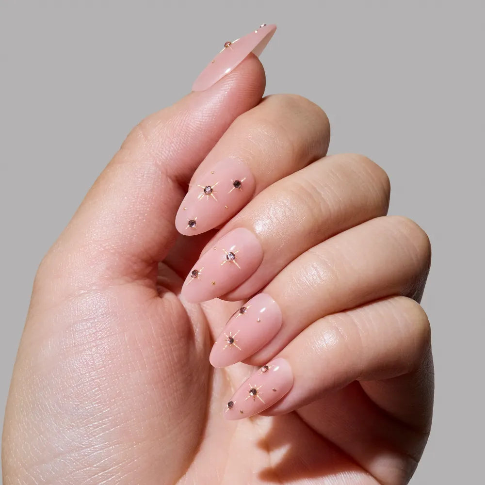 A hand with short almond nude pink nails displays the glamnetic Pink Comet set, decorated with gold foil stars and dots, against a plain gray background. - Product image