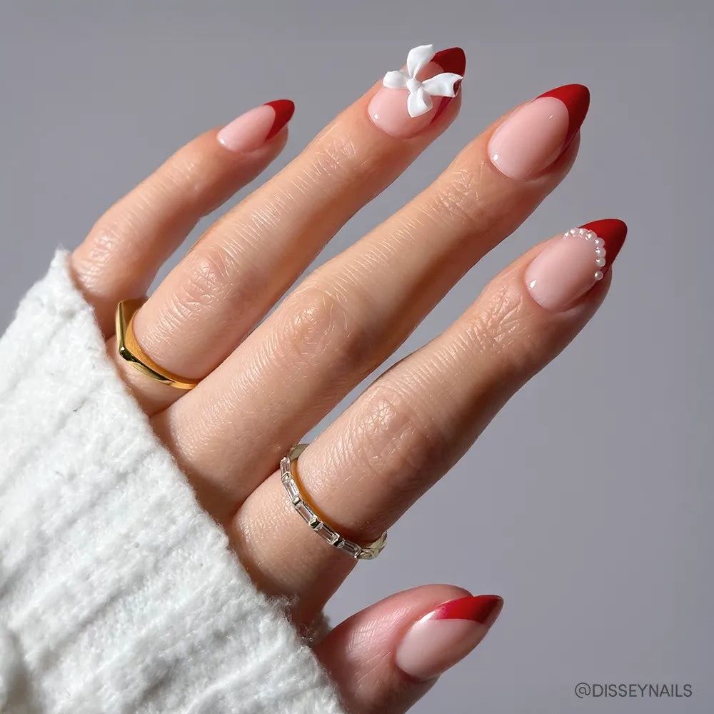 A hand with almond nails wears glamnetic's Ruby Ribbons: a pink base, bold ribbon-red tips, and a 3D white flower. One nail has tiny pearls. Gold rings and a white knit sleeve finish this chic holiday manicure.