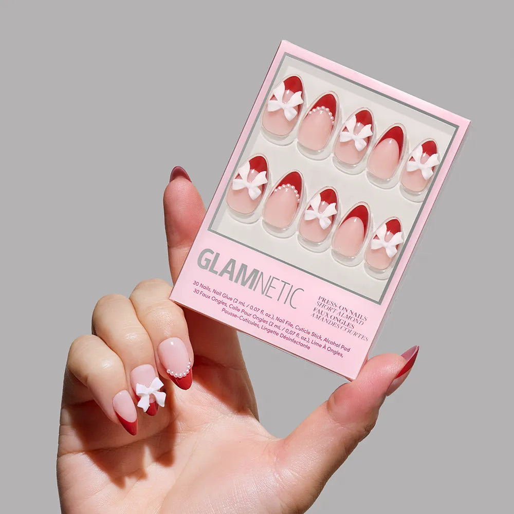 A hand with long, almond-shaped nails in pink and red, featuring a white 3D flower, holds a box of glamnetic's Ruby Ribbons press-on nails. Perfect for holiday manicures, the box displays ten matching nails.