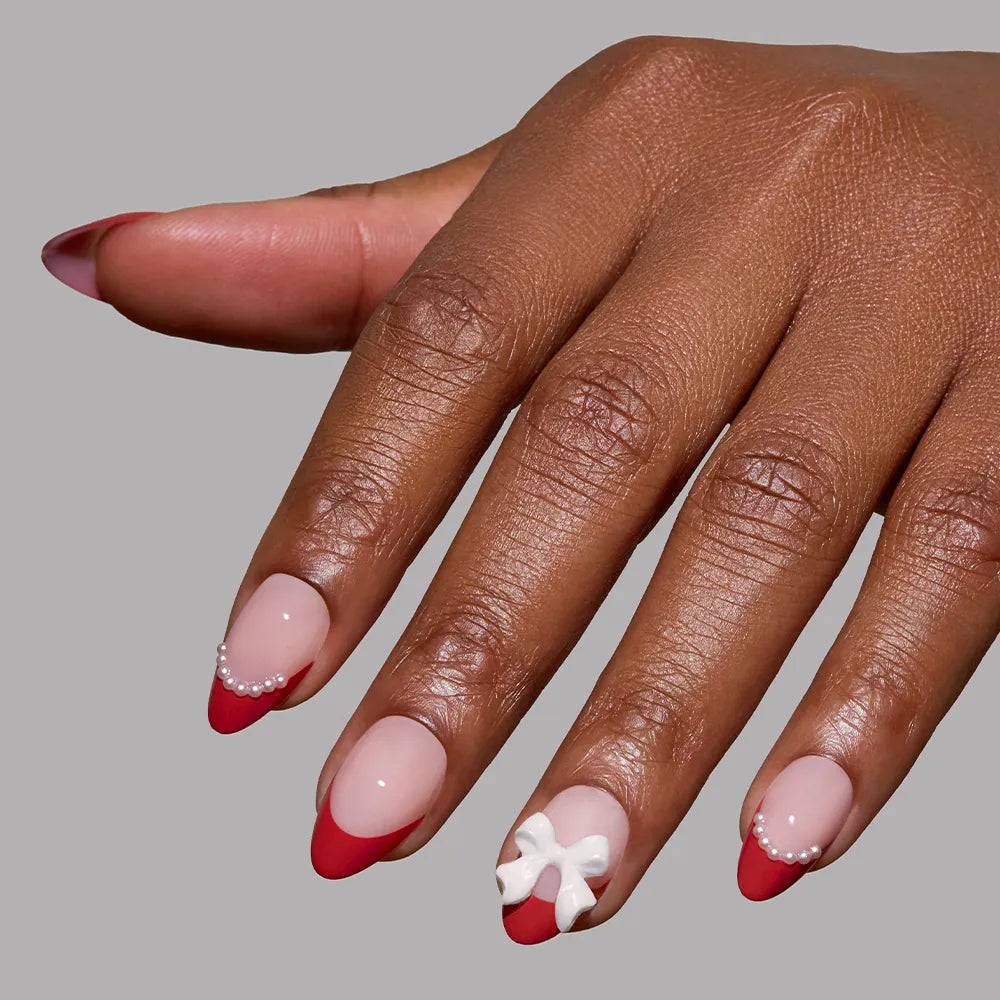A hand with almond nails features a holiday manicure using glamnetic’s Ruby Ribbons: red French tips, tiny white pearls on the ring and pinky nails, and a 3D white bow on the middle finger. The background is plain gray. - Product image
