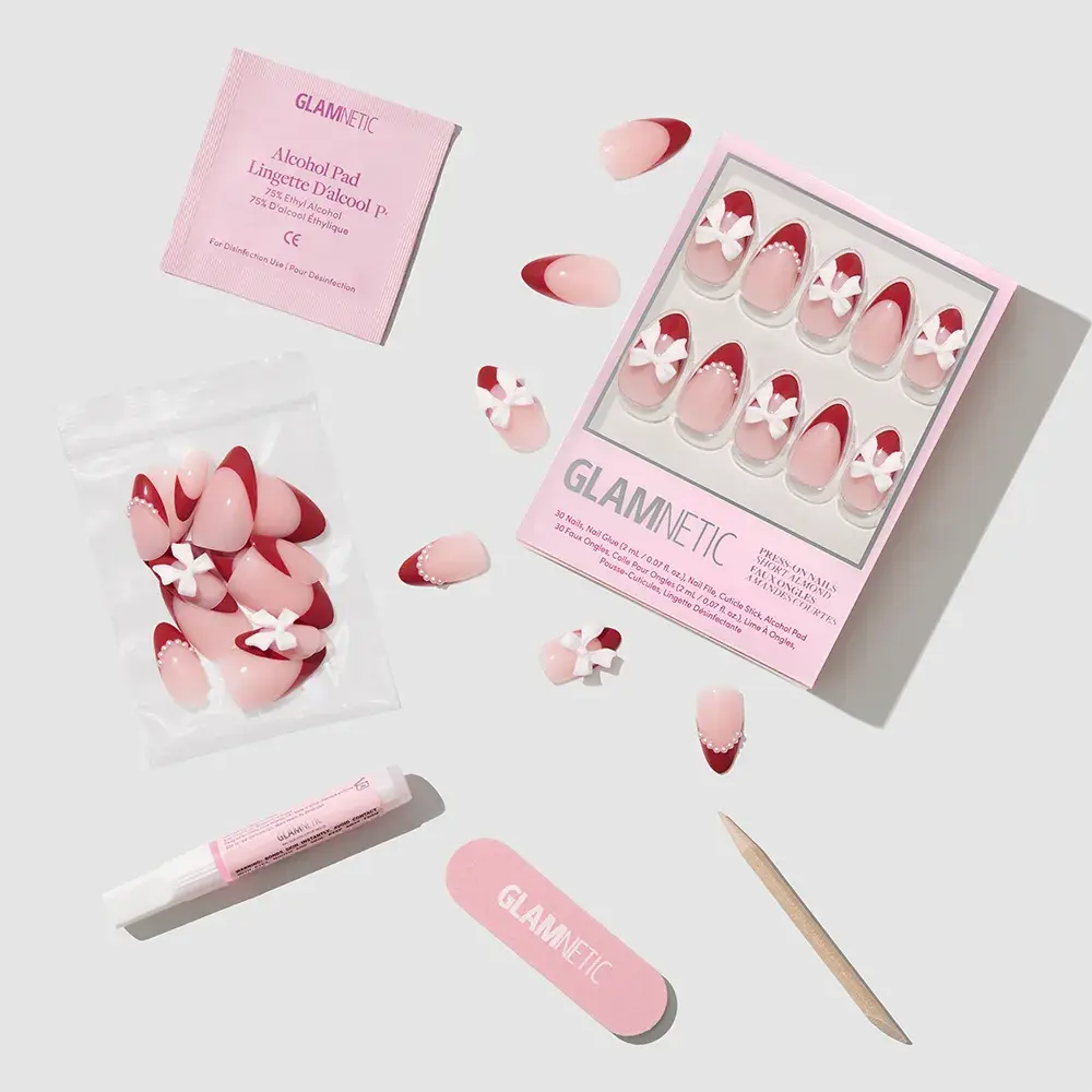 The Ruby Ribbons press-on nails kit by glamnetic features almond nails with pink, red, and white floral designs, perfect for holidays. Includes a nail file, cuticle stick, glue, alcohol pad, and extra nails, all neatly arranged on a light background. - Product image