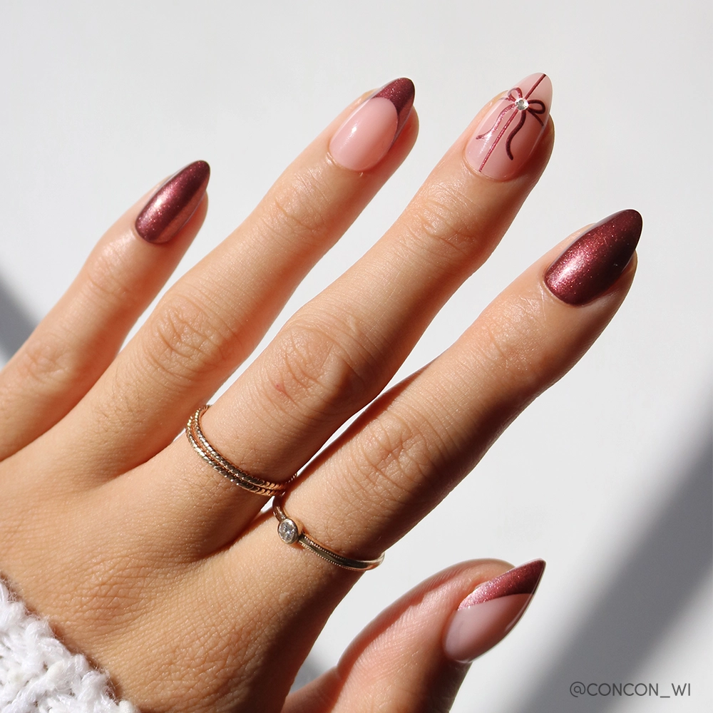 The "She's A Gift" nail set by glamnetic features short almond nails in metallic burgundy chrome and pink, with a bow and gem on the ring finger, plus gold rings on the index and middle fingers for an elegant look.