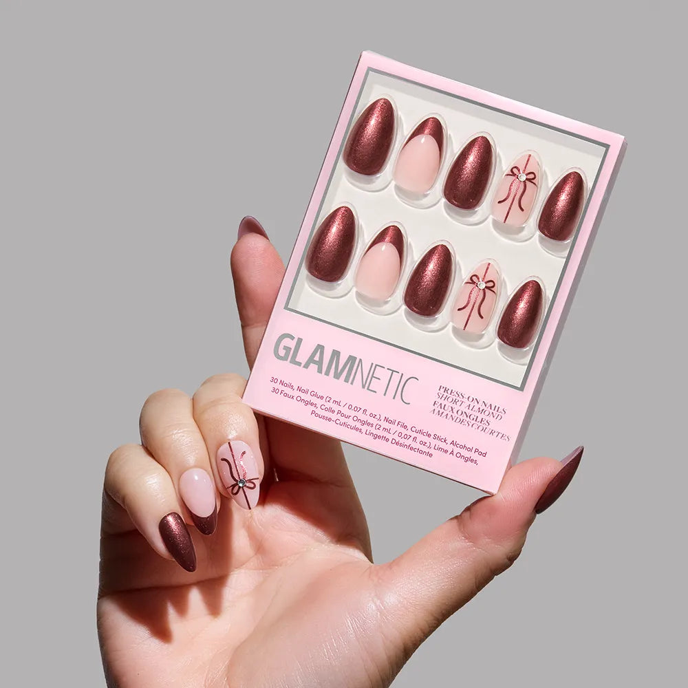 A hand holds glamnetic's She's A Gift nail set, showcasing short almond nails in metallic burgundy chrome with pale pink accent nails featuring delicate burgundy bow designs. The person’s own nails match the set’s elegant style.