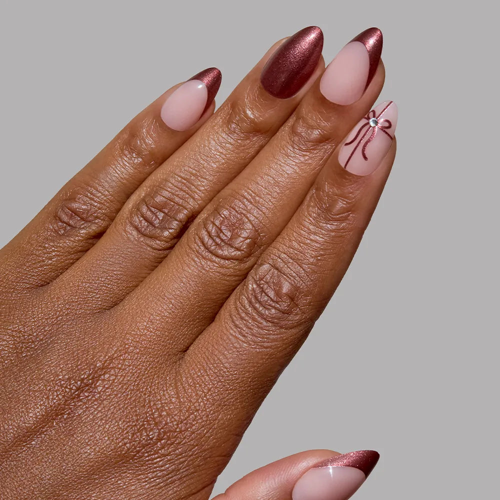 A hand with short almond nails wears glamnetic’s "She’s A Gift" set—burgundy chrome French tips with a matte pink base, plus a delicate bow accent nail. The background is plain gray. - Product image