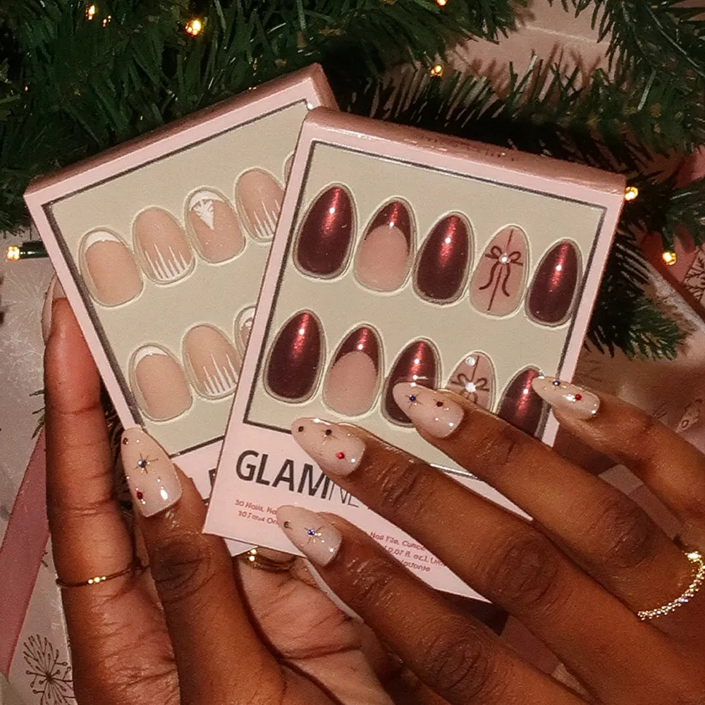 A person with festive short almond nails holds two glamnetic "She's A Gift" press-on nail sets—one with nude and white designs, the other featuring glossy burgundy chrome and gift accents—by a decorated Christmas tree. - Product image
