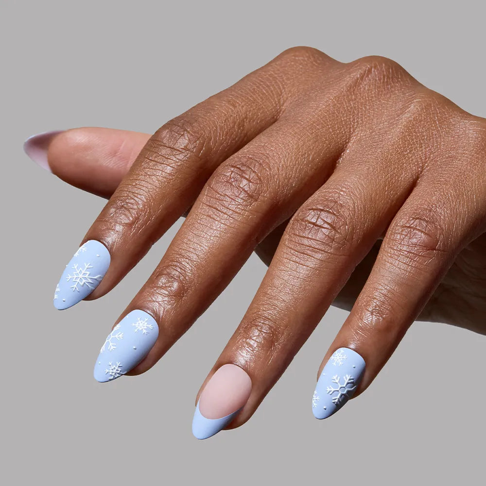 A hand with long matte almond Snow Bunny nails by glamnetic, painted light blue with white snowflakes and one nude half-moon accent, shown against a plain gray background for the perfect winter look. - Product image